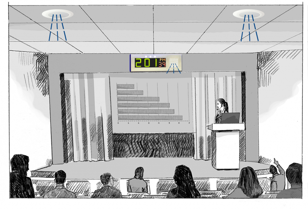 A woman with a headset microphone presents a bar chart in a lecture hall, while IP devices amplify her voice.